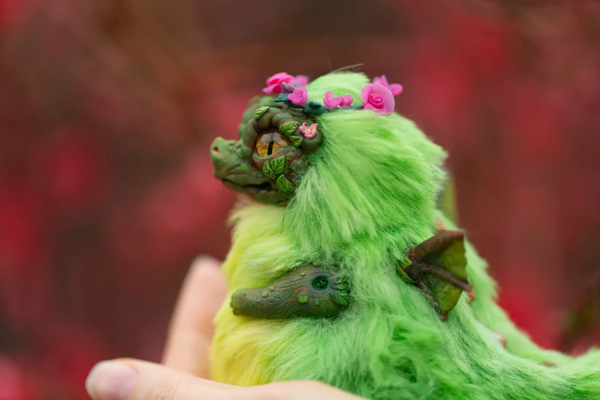 Кукла дракон сидит в человеческой руке. На голове у дракона венок из розовых цветов. Венок выполнен из полимерной глины. У дракончика на спине есть зеленые крылышки небольшого размера. Сам дракончик размером с маленького голубя.